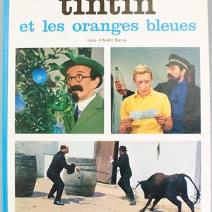 Puede incluir: Portada del libro "Tint&iacute;n y las naranjas azules". El t&iacute;tulo est&aacute; en letras negras grandes sobre un t&iacute;tulo azul. La portada presenta im&aacute;genes de personajes y escenas de la pel&iacute;cula, con el nombre del editor, Casterman, en la parte inferior.