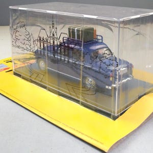 May include: A blue toy car with luggage on top, displayed in a clear plastic case. The case has a drawing of a building and trees. The car sits on a yellow cardboard base with text.