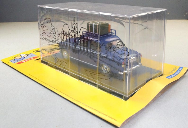 May include: A blue toy car with luggage on top, displayed in a clear plastic case. The case has a drawing of a building and trees. The car sits on a yellow cardboard base with text.