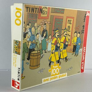 Peut inclure: Un puzzle de 100 pi&egrave;ces repr&eacute;sentant une sc&egrave;ne de la bande dessin&eacute;e Tintin. Le puzzle repr&eacute;sente Tintin et ses compagnons dans un march&eacute; de rue chinois. La bo&icirc;te est jaune et bleue avec le texte "Tintin" et "Puzzle 100" imprim&eacute; dessus.