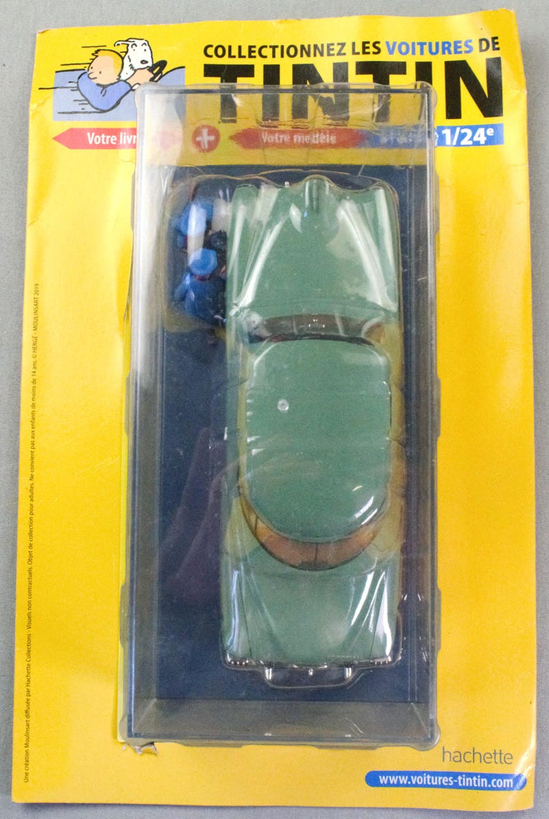 Pu&ograve; includere: Modello di auto verde vintage in una custodia di plastica trasparente, parte della collezione "Tintin". La confezione &egrave; gialla con testo francese e il sito web www.voitures-tintin.com.