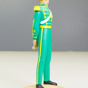 May include: A figurine of a man in a green military-style uniform with yellow accents. The figure has blonde hair and black shoes. The uniform includes a white sash and epaulettes. The figurine stands on a beige circular base.