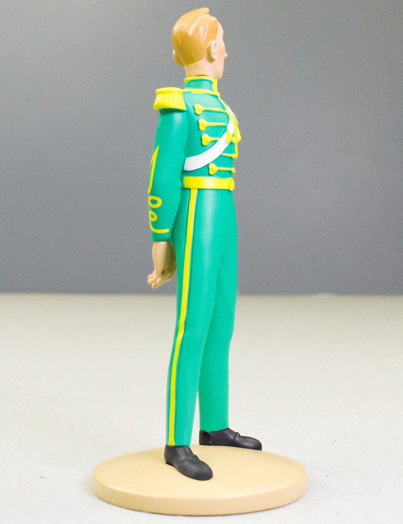 May include: A figurine of a man in a green military-style uniform with yellow accents. The figure has blonde hair and black shoes. The uniform includes a white sash and epaulettes. The figurine stands on a beige circular base.
