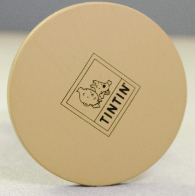 May include: A circular, light beige disc featuring a black line-art graphic of a character and a dog inside a square frame. The text TINTIN is printed in black capital letters within the frame, positioned diagonally across the center of the disc.