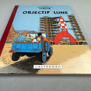 May include: A hardcover comic book titled "OBJECTIF LUNE" featuring a blue jeep with passengers, a red and white rocket, and a construction site. The cover has a light blue background with the text "HERGE LES AVENTURES DE TINTIN". The publisher's name, "CASTERMAN", is also visible.