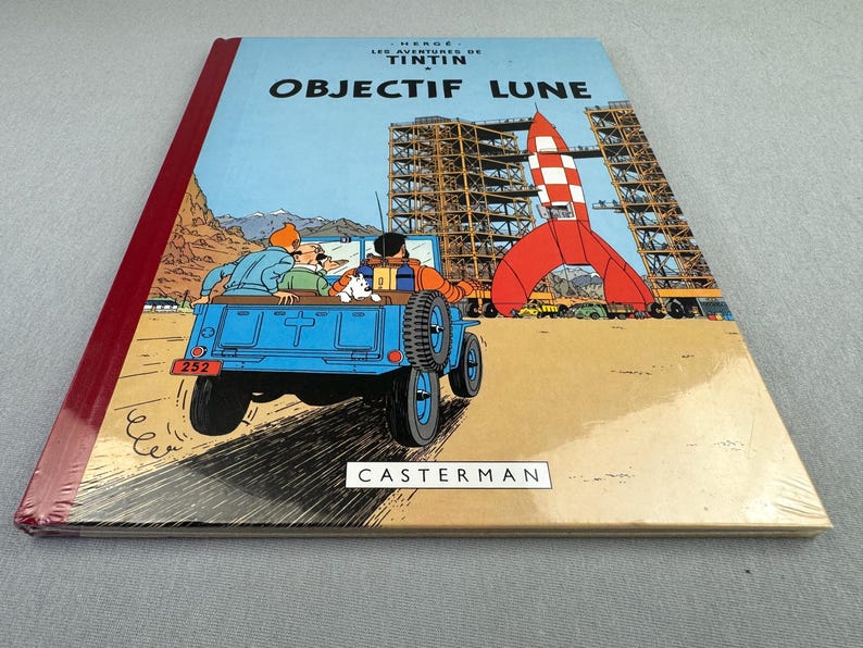 May include: A hardcover comic book titled "OBJECTIF LUNE" featuring a blue jeep with passengers, a red and white rocket, and a construction site. The cover has a light blue background with the text "HERGE LES AVENTURES DE TINTIN". The publisher's name, "CASTERMAN", is also visible.
