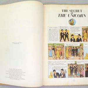 May include: Open book with the title "THE SECRET OF THE UNICORN" and comic strip illustrations. The pages are cream-colored with text and panels of drawings. The book is open to a double-page spread.