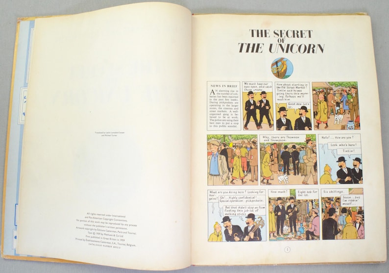 May include: Open book with the title "THE SECRET OF THE UNICORN" and comic strip illustrations. The pages are cream-colored with text and panels of drawings. The book is open to a double-page spread.