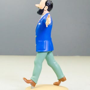 以下が含まれることがあります： 青いジャケット、緑のズボン、茶色の靴を履いた男性のフィギュア。フィギュアは黒いひげと髪を持ち、ベージュ色の台座を歩いています。フィギュアは右腕を伸ばしています。