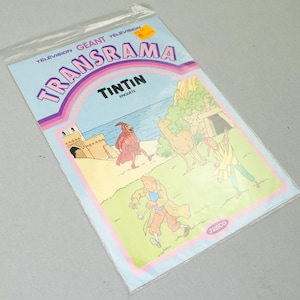 K&ouml;nnte beinhalten: Ein Vintage-Comic "Transrama" mit Tim und Struppi in einer Plastikh&uuml;lle. Das Cover ist hellblau mit Illustrationen von Figuren und Geb&auml;uden. Der Text enth&auml;lt "Tintin Enqu&ecirc;te" und "Television Geant Transrama".