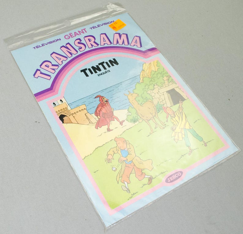 K&ouml;nnte beinhalten: Ein Vintage-Comic "Transrama" mit Tim und Struppi in einer Plastikh&uuml;lle. Das Cover ist hellblau mit Illustrationen von Figuren und Geb&auml;uden. Der Text enth&auml;lt "Tintin Enqu&ecirc;te" und "Television Geant Transrama".