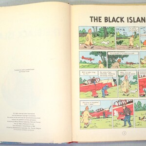 K&ouml;nnte beinhalten: Offenes Buch mit einem Comic-Strip mit dem Titel "Die schwarze Insel". Die Comic-Panels zeigen einen Mann in einem gelben Mantel, einen wei&szlig;en Hund und ein rotes Flugzeug. Sprechblasen enthalten Dialoge.
