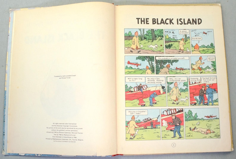 K&ouml;nnte beinhalten: Offenes Buch mit einem Comic-Strip mit dem Titel "Die schwarze Insel". Die Comic-Panels zeigen einen Mann in einem gelben Mantel, einen wei&szlig;en Hund und ein rotes Flugzeug. Sprechblasen enthalten Dialoge.