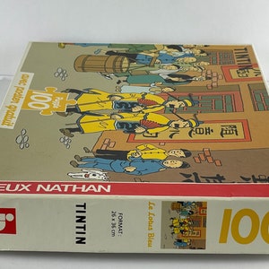 Peut inclure: Bo&icirc;te de puzzle de 100 pi&egrave;ces repr&eacute;sentant Tintin et des personnages de la bande dessin&eacute;e "Le Lotus Bleu". La bo&icirc;te est rouge et blanche avec une image de Tintin et des personnages sur le devant. La bo&icirc;te comprend &eacute;galement le texte "Tintin" et "Le Lotus Bleu".
