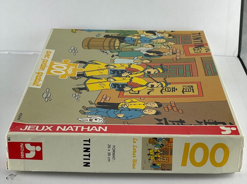 Peut inclure: Bo&icirc;te de puzzle de 100 pi&egrave;ces repr&eacute;sentant Tintin et des personnages de la bande dessin&eacute;e "Le Lotus Bleu". La bo&icirc;te est rouge et blanche avec une image de Tintin et des personnages sur le devant. La bo&icirc;te comprend &eacute;galement le texte "Tintin" et "Le Lotus Bleu".