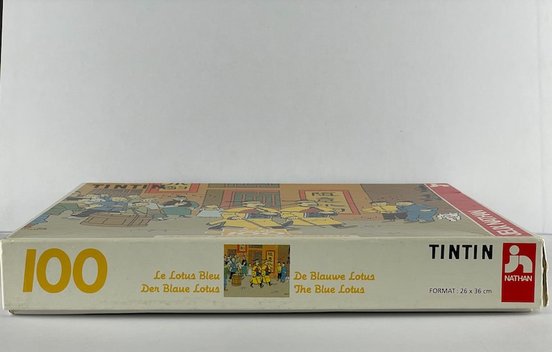 Peut inclure: Une bo&icirc;te blanche avec une bordure jaune et le nombre 100 imprim&eacute; en jaune. La bo&icirc;te contient un puzzle de 100 pi&egrave;ces du livre de bande dessin&eacute;e de Tintin "Le Lotus Bleu". La bo&icirc;te comprend &eacute;galement le titre de la bande dessin&eacute;e en fran&ccedil;ais, allemand, n&eacute;erlandais et anglais.