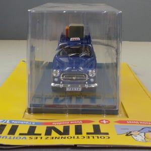 May include: A blue toy taxi model with luggage on the roof, displayed in a clear plastic case. The model car has a white "TAXI" sign and a license plate. The packaging is yellow with black text.