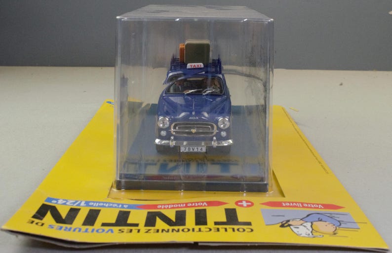 May include: A blue toy taxi model with luggage on the roof, displayed in a clear plastic case. The model car has a white "TAXI" sign and a license plate. The packaging is yellow with black text.