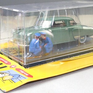 Pu&ograve; includere: Modello di auto d'epoca verde con due figure in miniatura in uniforme blu, racchiuso in una vetrina di plastica trasparente. Il modello &egrave; posto su una base di cartone gialla con testo francese e illustrazione.
