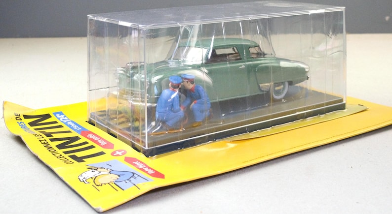 Pu&ograve; includere: Modello di auto d'epoca verde con due figure in miniatura in uniforme blu, racchiuso in una vetrina di plastica trasparente. Il modello &egrave; posto su una base di cartone gialla con testo francese e illustrazione.