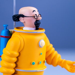 May include: A yellow plastic figurine of a man wearing a yellow diving suit with a blue oxygen tank on his back. The man has a bald head, a black mustache, and is wearing glasses.