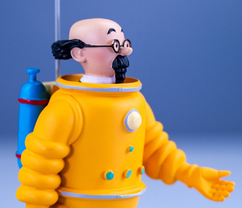 May include: A yellow plastic figurine of a man wearing a yellow diving suit with a blue oxygen tank on his back. The man has a bald head, a black mustache, and is wearing glasses.