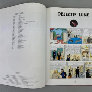 Puede incluir: Libro abierto con el t&iacute;tulo "OBJECTIF LUNE" e ilustraciones de c&oacute;mic. La p&aacute;gina izquierda contiene texto. La p&aacute;gina derecha presenta una tira c&oacute;mica con un cohete y un coche azul.