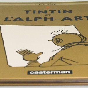 Może przedstawiać: Książka w twardej oprawie zatytułowana "TINTIN et L'ALPH-ART" autorstwa Herg&eacute;. Okładka jest złota z czarnym tekstem i rysunkiem Tintina. Grzbiet jest pomarańczowy, a logo wydawcy, "casterman", znajduje się na dole.