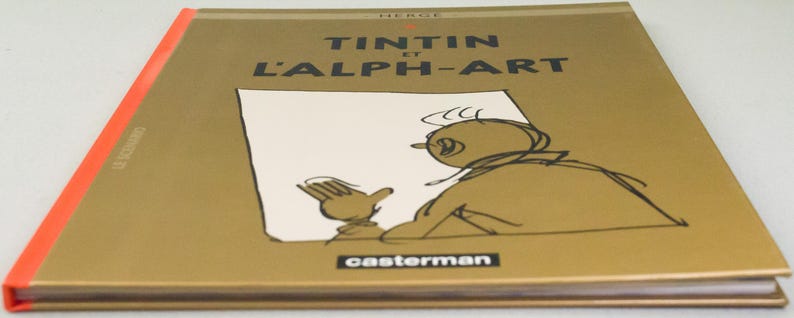 Może przedstawiać: Książka w twardej oprawie zatytułowana "TINTIN et L'ALPH-ART" autorstwa Herg&eacute;. Okładka jest złota z czarnym tekstem i rysunkiem Tintina. Grzbiet jest pomarańczowy, a logo wydawcy, "casterman", znajduje się na dole.