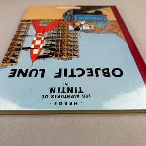 May include: A hardcover book titled "OBJECTIF LUNE" by Herg&eacute;. The cover features a rocket, a construction, and a blue sky background. The book's spine is red. The text "LES AVENTURES DE TINTIN" is also visible.