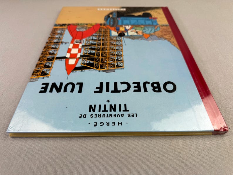 May include: A hardcover book titled "OBJECTIF LUNE" by Herg&eacute;. The cover features a rocket, a construction, and a blue sky background. The book's spine is red. The text "LES AVENTURES DE TINTIN" is also visible.