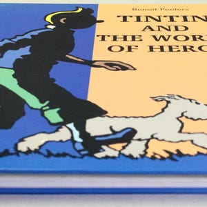 Puede incluir: Un libro de tapa dura titulado "Tint&iacute;n y el mundo de Herg&eacute;". La portada presenta un fondo dividido azul y naranja con una ilustraci&oacute;n de dibujos animados de Tint&iacute;n y su perro corriendo. El lomo del libro es azul.