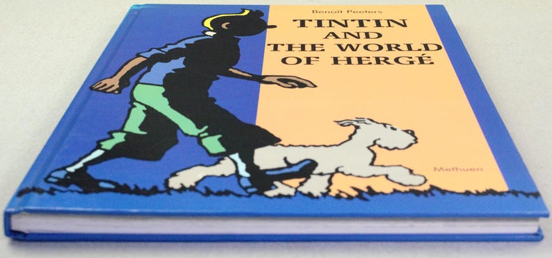 Puede incluir: Un libro de tapa dura titulado "Tint&iacute;n y el mundo de Herg&eacute;". La portada presenta un fondo dividido azul y naranja con una ilustraci&oacute;n de dibujos animados de Tint&iacute;n y su perro corriendo. El lomo del libro es azul.