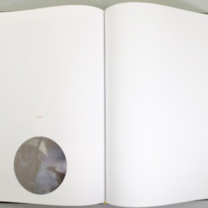 May include: An open book with blank white pages. A circular image is embedded on the left page. The book has a black spine, and the page edges are visible. The right page is also blank.