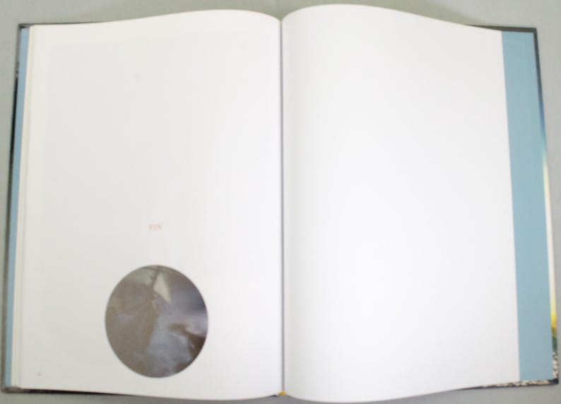 May include: An open book with blank white pages. A circular image is embedded on the left page. The book has a black spine, and the page edges are visible. The right page is also blank.