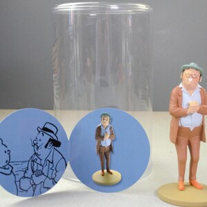 May include: A figurine of a man wearing a tan suit, white shirt, and green hat, standing on a circular base. Beside it are two circular cards featuring a line drawing of a man and a boy, and a photograph of the same figurine in a clear container.