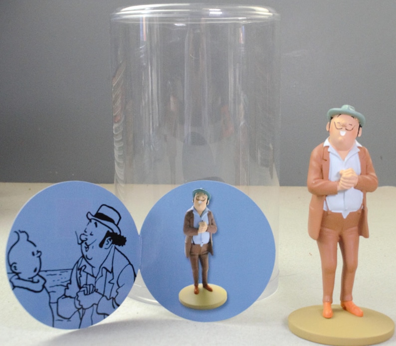 May include: A figurine of a man wearing a tan suit, white shirt, and green hat, standing on a circular base. Beside it are two circular cards featuring a line drawing of a man and a boy, and a photograph of the same figurine in a clear container.