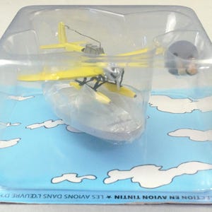 Op de afbeelding: Een geel watervliegtuigmodel in een doorzichtige plastic verpakking. Het vliegtuig heeft een zilveren propeller en zwarte details. De verpakking staat op een blauwe achtergrond met witte wolkenillustraties. De verpakking is gelabeld "COLLECTION EN AVION TINTIN".