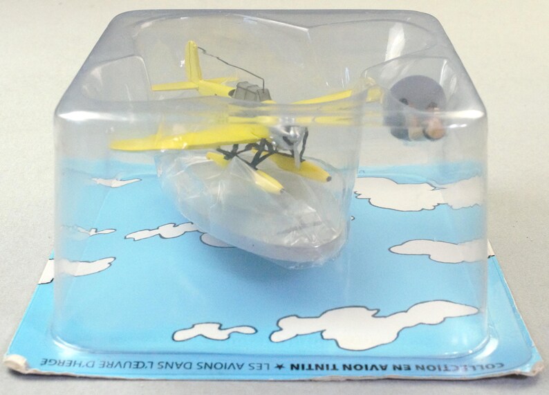 Op de afbeelding: Een geel watervliegtuigmodel in een doorzichtige plastic verpakking. Het vliegtuig heeft een zilveren propeller en zwarte details. De verpakking staat op een blauwe achtergrond met witte wolkenillustraties. De verpakking is gelabeld "COLLECTION EN AVION TINTIN".