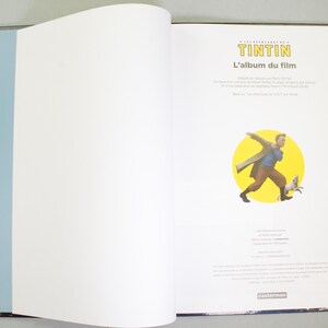 May include: An open book with a blue spine and a black border. The right page features a cartoon illustration of Tintin and Snowy. The book's title is "Tintin: L'album du film."