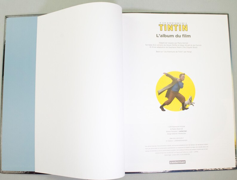 May include: An open book with a blue spine and a black border. The right page features a cartoon illustration of Tintin and Snowy. The book's title is "Tintin: L'album du film."