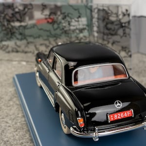 Puede incluir: Un modelo de coche Mercedes-Benz negro con un acabado brillante. El coche tiene una matr&iacute;cula roja con texto blanco y un parachoques cromado. El modelo est&aacute; sobre una base azul. El interior del coche es visible a trav&eacute;s de las ventanas.