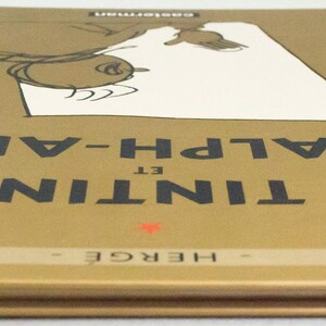 Może przedstawiać: Twarda oprawa książki zatytułowanej "Tintin et l'Alph-Art" autorstwa Herg&eacute;. Okładka jest złota z czarnym tekstem i ilustracją z kresk&oacute;wek. Grzbiet jest pomarańczowy. Wydane przez Casterman.