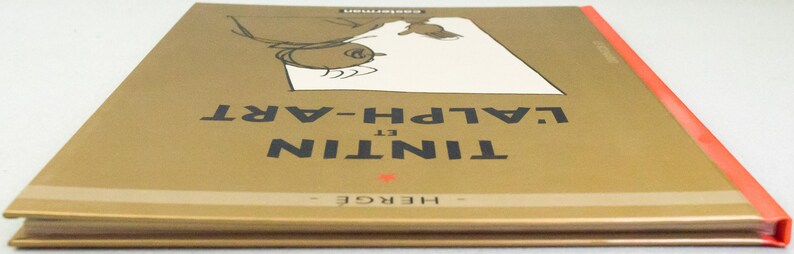 Może przedstawiać: Twarda oprawa książki zatytułowanej "Tintin et l'Alph-Art" autorstwa Herg&eacute;. Okładka jest złota z czarnym tekstem i ilustracją z kresk&oacute;wek. Grzbiet jest pomarańczowy. Wydane przez Casterman.