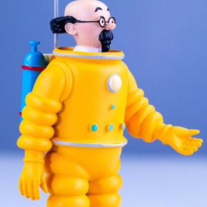 May include: A yellow astronaut figurine with a blue oxygen tank, black hair, glasses, and a mustache. The figurine is wearing a yellow spacesuit with a white collar and blue accents.