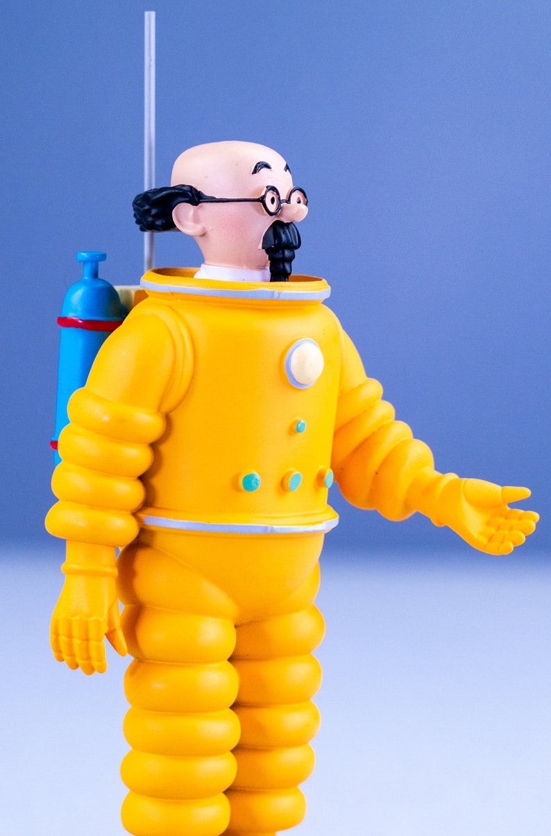 May include: A yellow astronaut figurine with a blue oxygen tank, black hair, glasses, and a mustache. The figurine is wearing a yellow spacesuit with a white collar and blue accents.