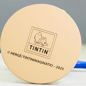 以下が含まれることがあります： タンタンとスノーウィーのタンタンのロゴが入った、薄いベージュ色のフィギュアの円形ベース。ベースには「&copy; HERG&Eacute;/TINTINIMAGINATIO - 2025」の文字が入っています。フィギュアには青とタンの腕があります。