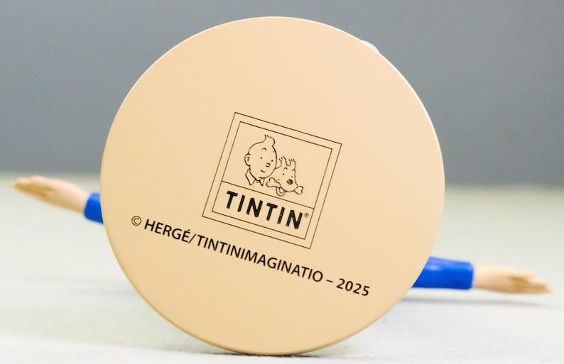 以下が含まれることがあります： タンタンとスノーウィーのタンタンのロゴが入った、薄いベージュ色のフィギュアの円形ベース。ベースには「&copy; HERG&Eacute;/TINTINIMAGINATIO - 2025」の文字が入っています。フィギュアには青とタンの腕があります。