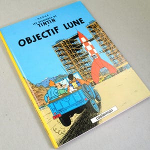 K&ouml;nnte beinhalten: Ein gebundenes Comicbuch mit dem Titel "OBJECTIF LUNE" auf Franz&ouml;sisch. Das Cover zeigt eine Rakete, einen blauen Jeep und Figuren vor blauem Himmel. Das Buch hat einen gelben Buchr&uuml;cken und der Verlag Casterman ist sichtbar.