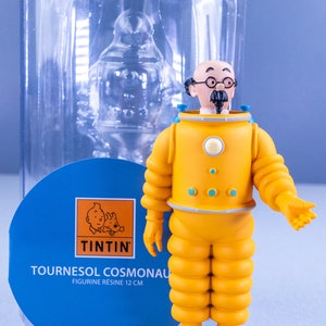 May include: A yellow and blue resin figurine of Professor Calculus from the Tintin comics, wearing a yellow space suit with a blue helmet. The figurine is 12 cm tall and is standing on a brown base. The figurine is packaged in a clear plastic container with a blue and white label that reads "Tournesol Cosmonaut Figurine Resine 12 cm" and features the Tintin logo.